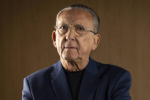 Retrato de Galvão Bueno, narrador esportivo, usando óculos e paletó azul escuro. Ele aparece sério em frente a um fundo neutro, em tom amadeirado.