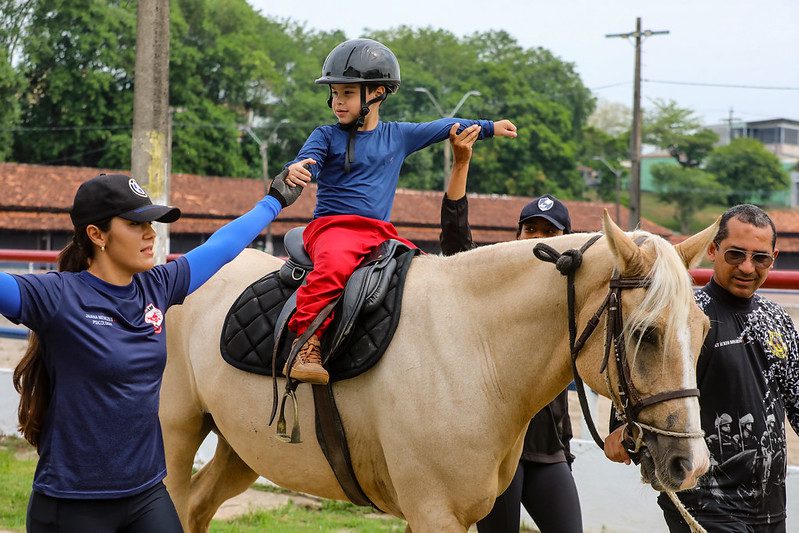 Criança com deficiência montada em cavalo durante atividade de equoterapia, acompanhada por equipe da Polícia Militar em Manaus.