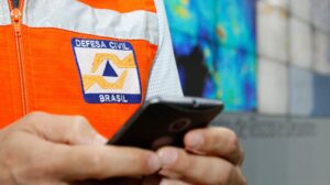 Agente da Defesa Civil Brasil com colete laranja utiliza celular para operação de alertas