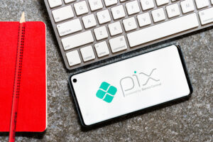 Smartphone exibindo a logo do Pix ao lado de teclado e caderno vermelho.