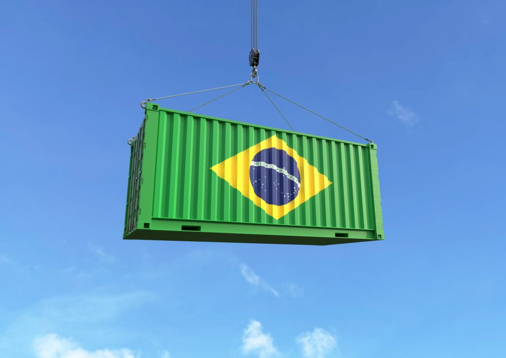Contêiner de carga verde com a bandeira do Brasil sendo içado por guindaste contra o céu azul.