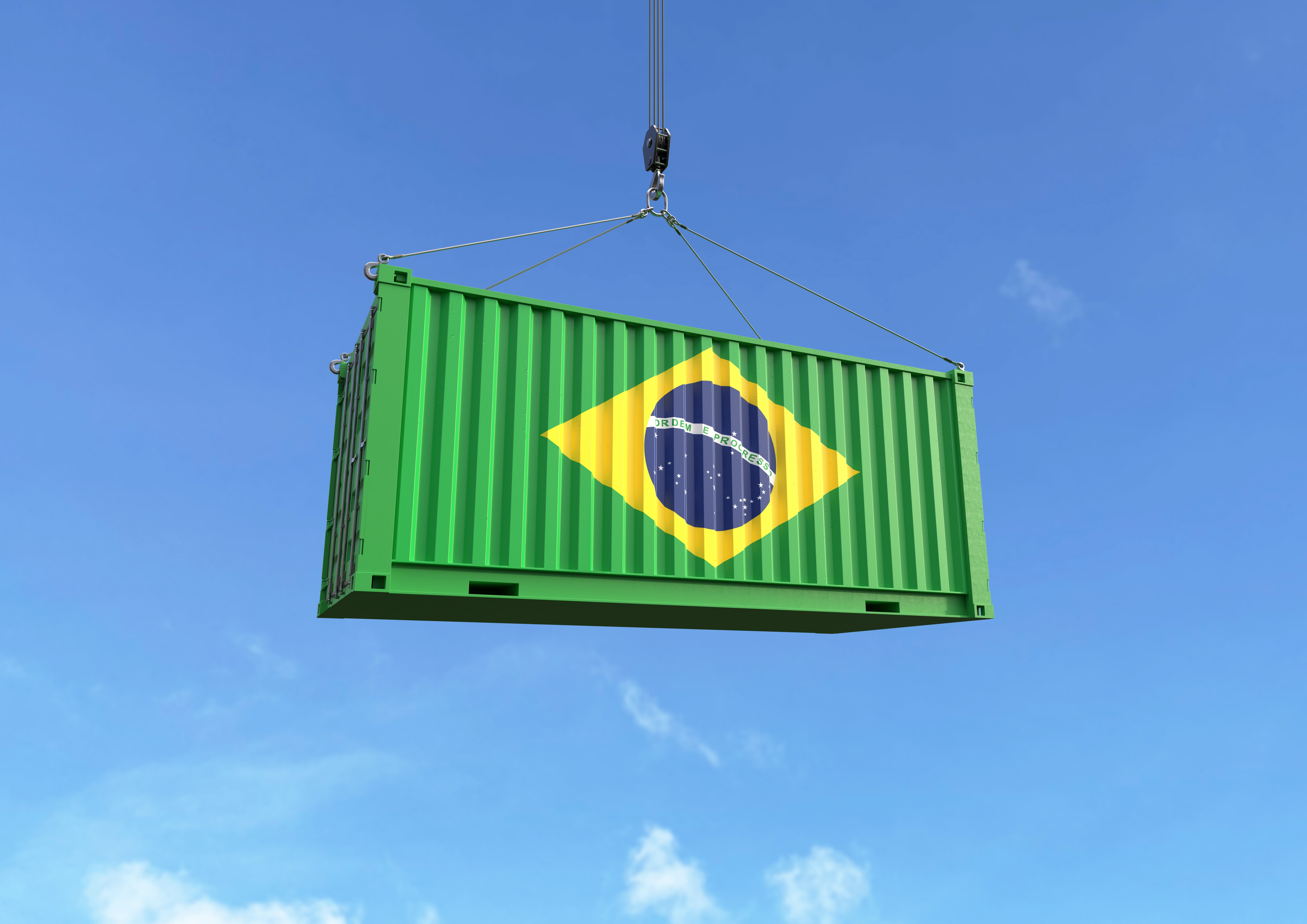 Contêiner de carga verde com a bandeira do Brasil sendo içado por guindaste contra o céu azul.