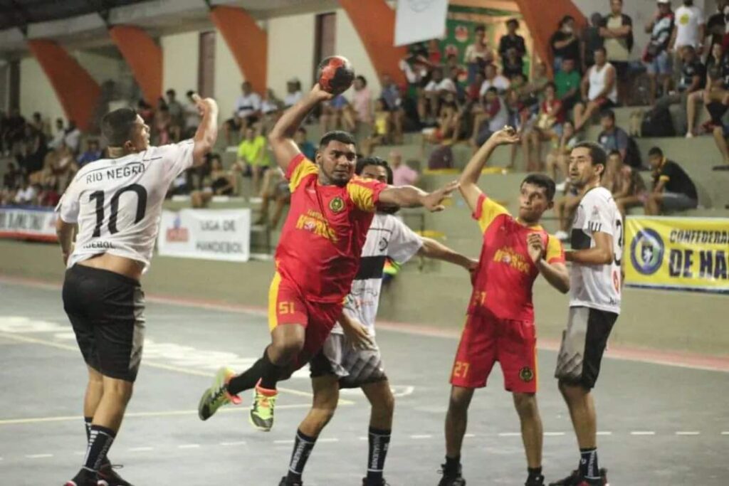 Atleta com uniforme vermelho realiza arremesso em partida de handebol contra equipe de uniforme branco durante o Campeonato Amazonense, no Ginásio do Rio Negro, em Manaus.