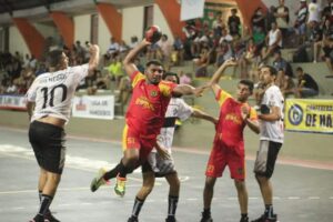 Atleta com uniforme vermelho realiza arremesso em partida de handebol contra equipe de uniforme branco durante o Campeonato Amazonense, no Ginásio do Rio Negro, em Manaus.