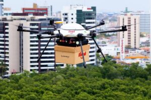 Drone de entrega do iFood em parceria com a Speedbird Aero transporta caixa sobre área urbana de Aracaju, com prédios residenciais e comerciais ao fundo.