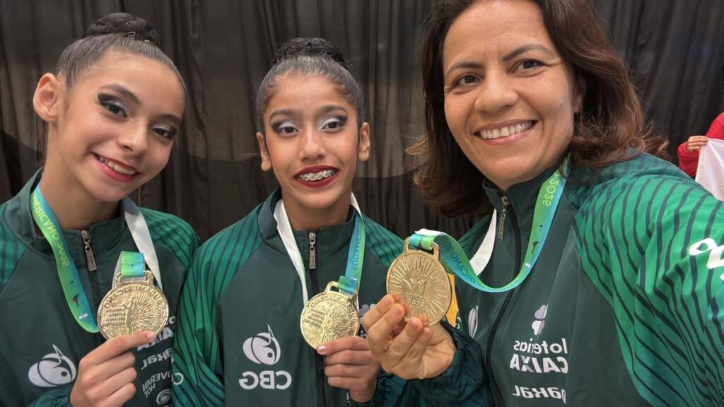 Duas ginastas adolescentes e sua técnica seguram medalhas de ouro e sorriem após vitória no Sul-Americano de Ginástica Rítmica, representando o Amazonas.