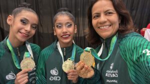 Duas ginastas adolescentes e sua técnica seguram medalhas de ouro e sorriem após vitória no Sul-Americano de Ginástica Rítmica, representando o Amazonas.