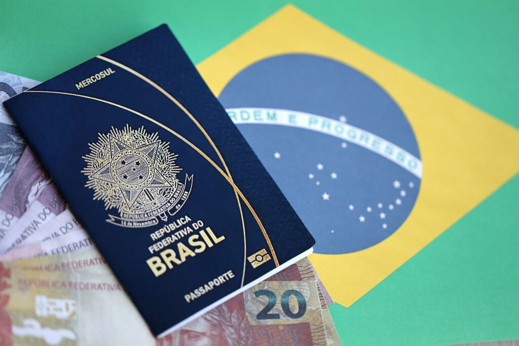 Passaporte da República Federativa do Brasil apoiado sobre notas de dinheiro e a bandeira nacional, representando o custo e a importância do documento para viagens internacionais.