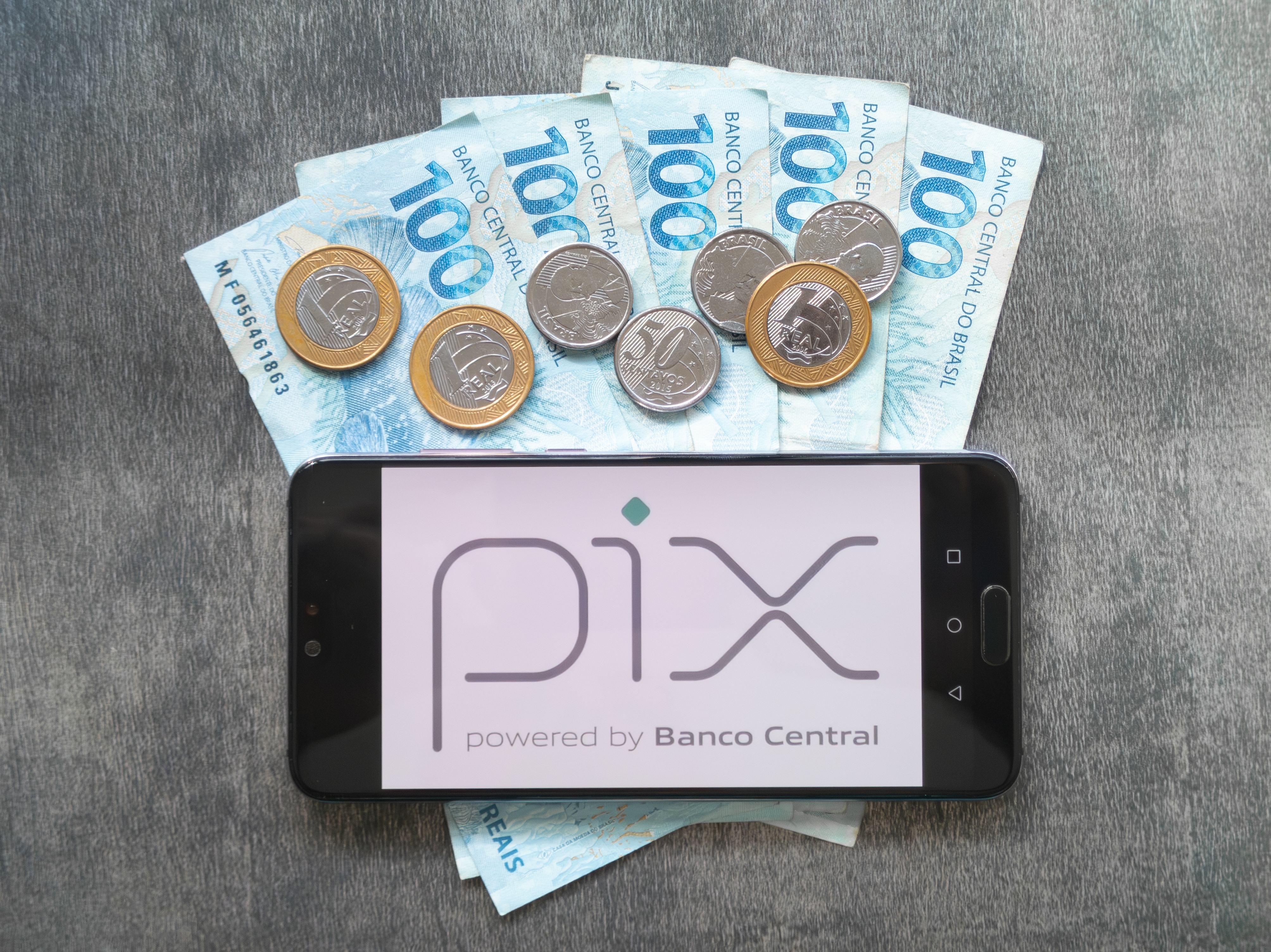 Celular com logo do Pix sobre notas e moedas brasileiras, simbolizando novo botão de contestação para fraudes.