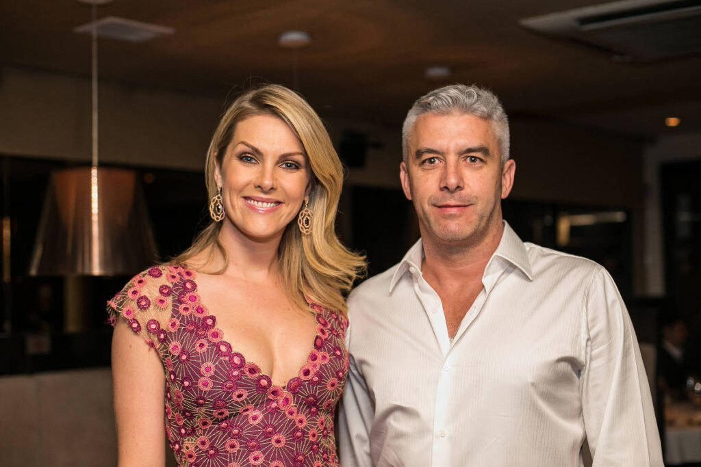 Ana Hickmann sorrindo ao lado de Alexandre Correa em um evento social; ela usa vestido rosa rendado, e ele veste camisa branca. Ambos posam para a câmera em um ambiente interno iluminado.