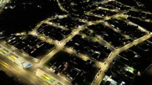 Imagem aérea noturna de um bairro com ruas iluminadas por lâmpadas LED, mostrando iluminação pública em funcionamento.