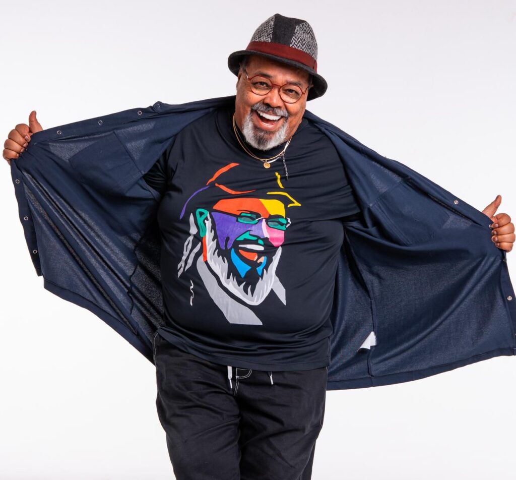 Jorge Aragão sorrindo, usando chapéu e camisa com arte colorida, em foto de divulgação do show no Porto de Manaus.