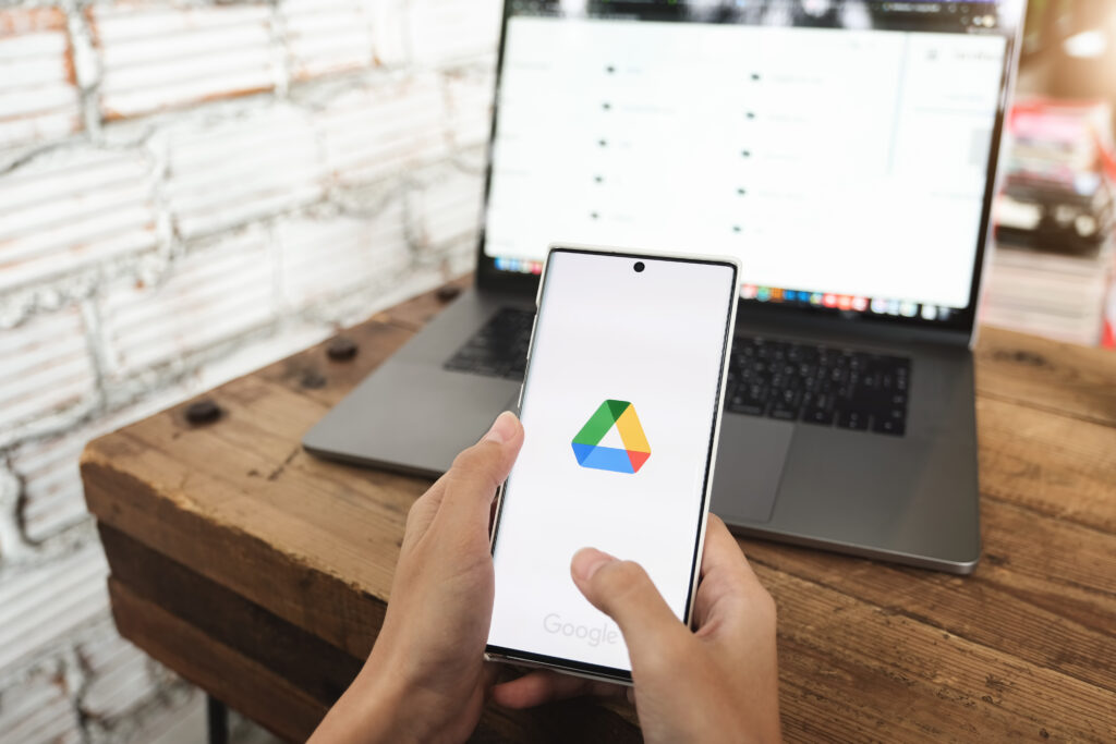 Homem segura celular com o Google Drive aberto enquanto visualiza o serviço na tela do computador.