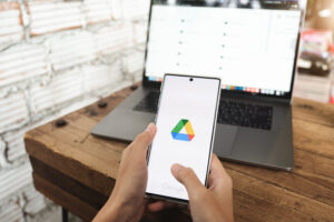 Homem segura celular com o Google Drive aberto enquanto visualiza o serviço na tela do computador.