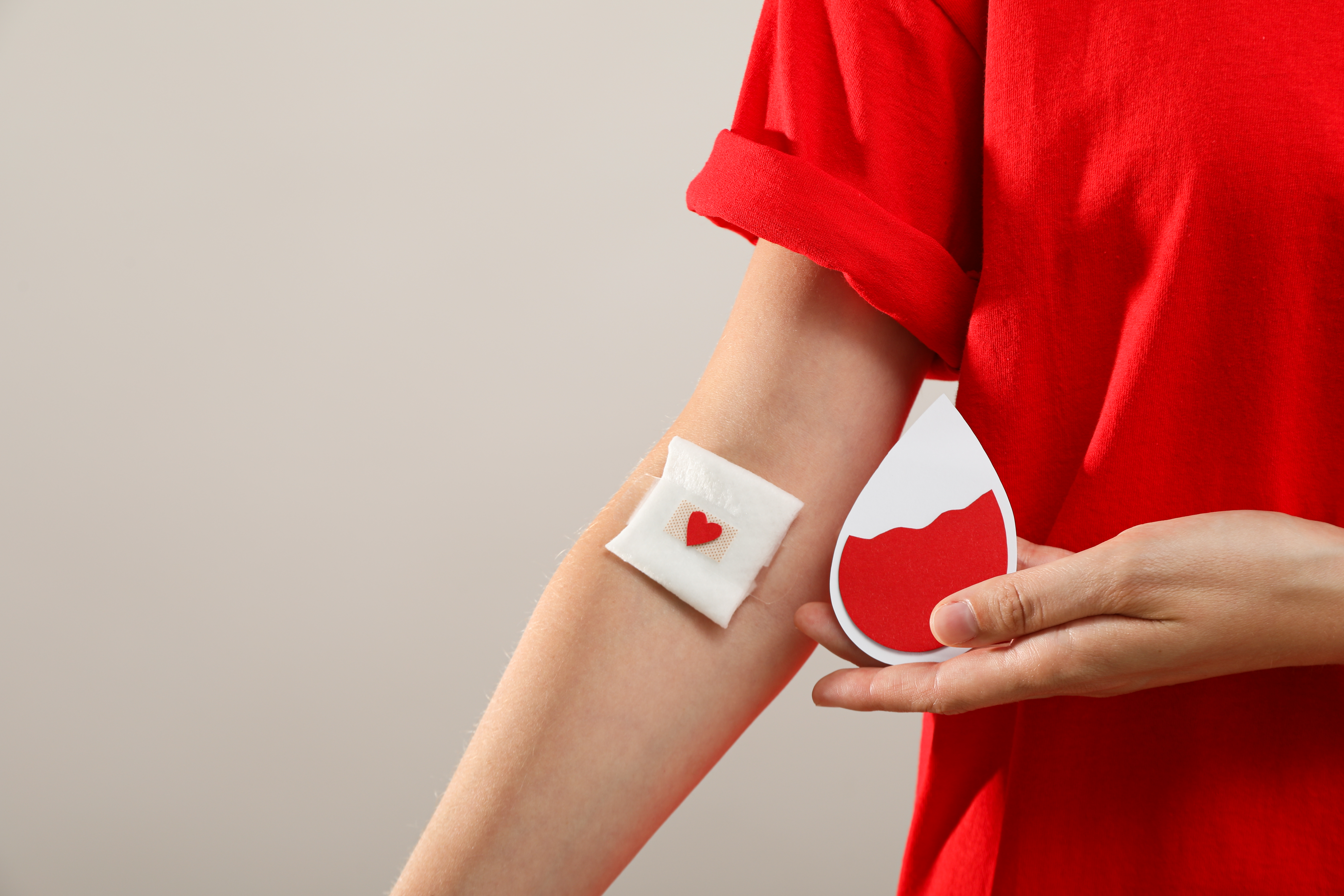 Braço com curativo após doação de sangue e mão segurando símbolo de gota vermelha, representando ação de doação.