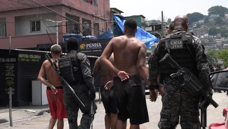 Policiais militares escoltam homens detidos em operação contra o crime organizado em comunidade urbana.