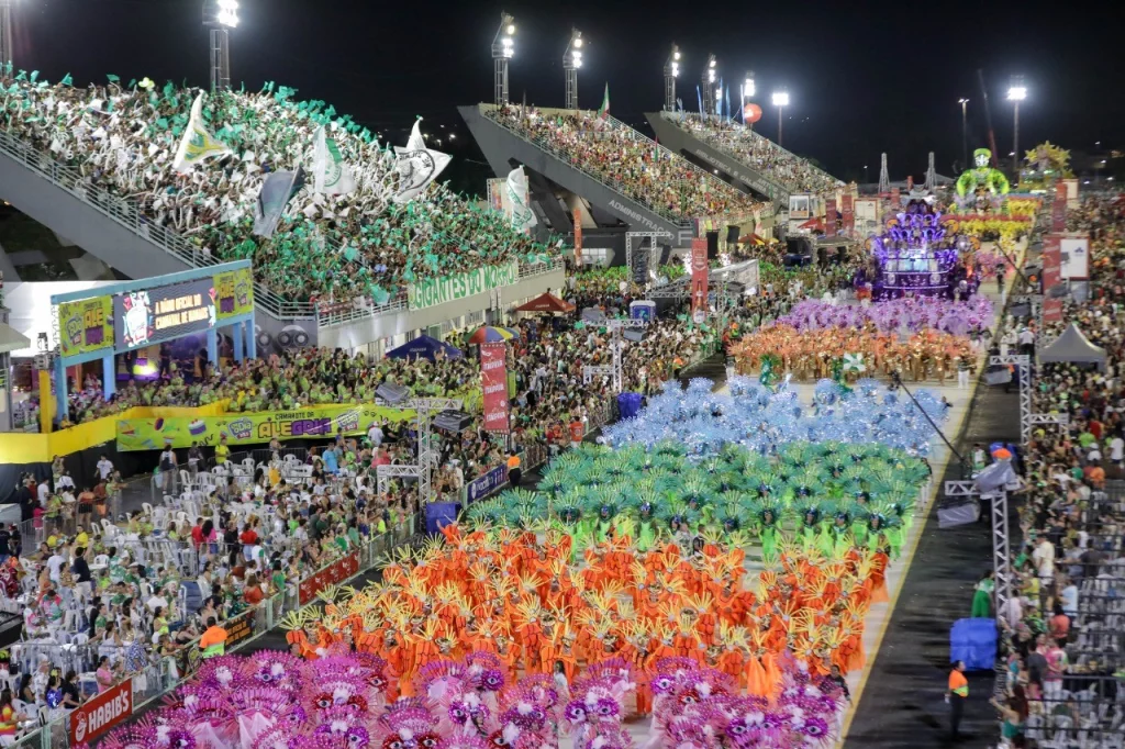 Vista aérea do desfile das escolas de samba de Manaus no Sambódromo, com arquibancadas cheias e alas coloridas ocupando a pista.