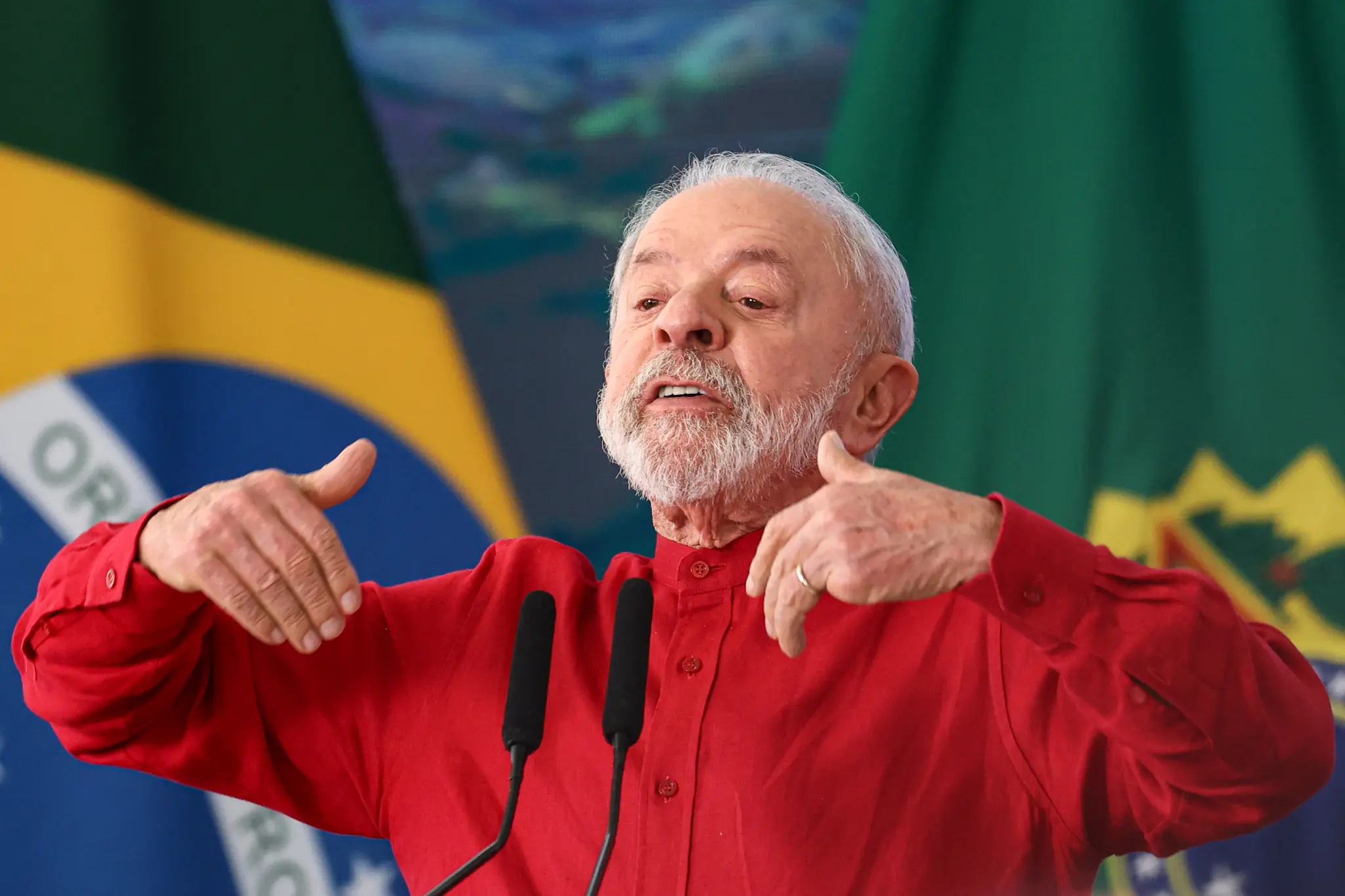 Presidente Luiz Inácio Lula da Silva gesticula durante discurso, com bandeira do Brasil ao fundo.