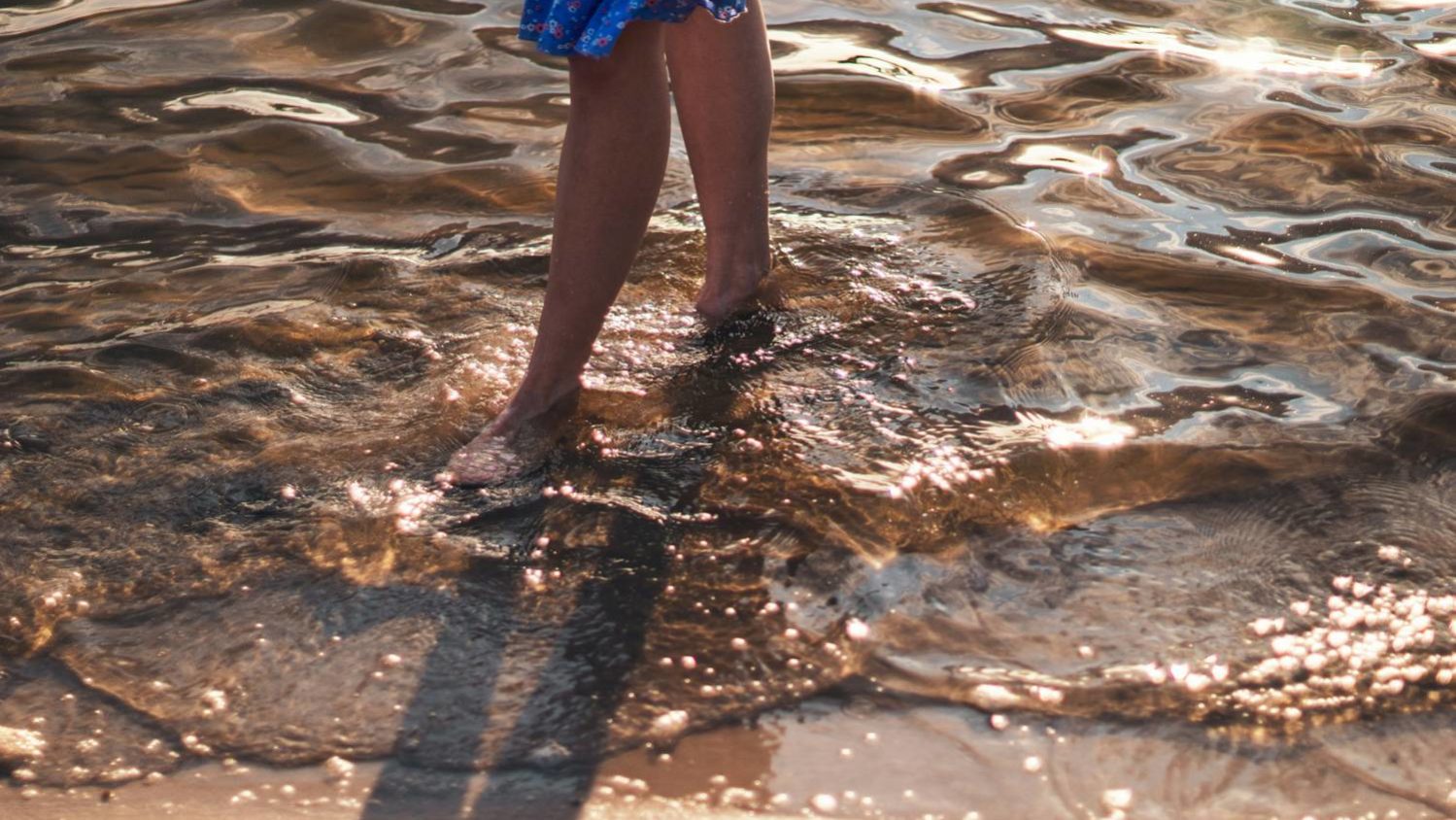 Pernas de uma pessoa caminhando na água rasa, com reflexos dourados do sol sobre a superfície do mar.