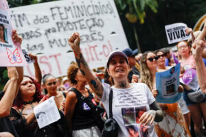 Mulheres protestam contra feminicídio segurando cartazes e levantando o braço durante ato nacional no Brasil.