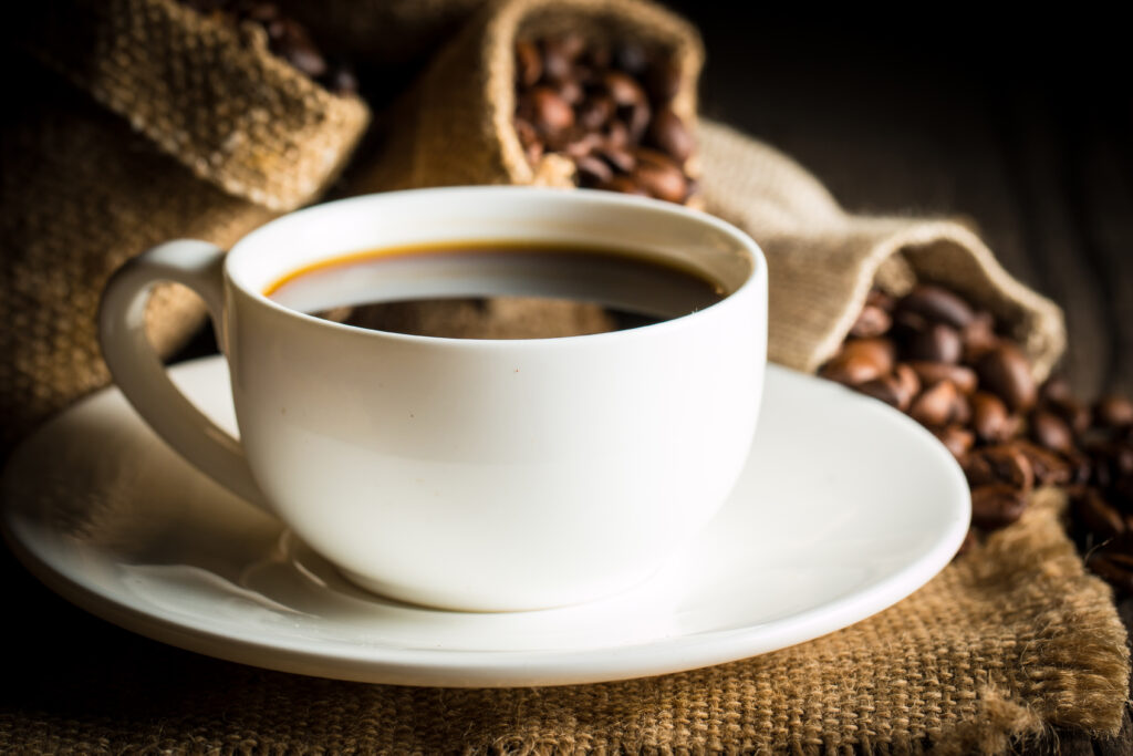 Xícara de café preto ao lado de grãos torrados sobre pano rústico, simbolizando estudo que associa o consumo moderado de café ao envelhecimento celular mais lento.