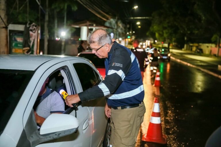 Agente realiza teste do bafômetro em motorista durante fiscalização de trânsito à noite, com cones de sinalização na via.