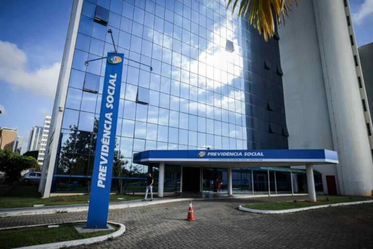 Fachada de prédio da Previdência Social com estrutura de vidro e placa azul de identificação na entrada.