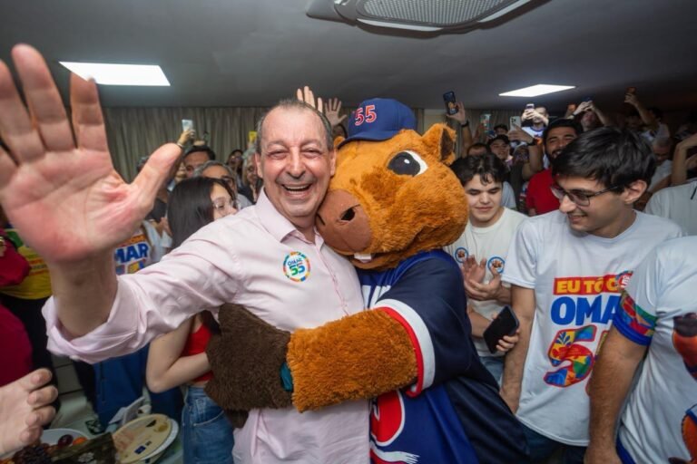 Omar Aziz sorri e acena para apoiadores enquanto abraça um mascote de sua campanha após a vitória nas eleições para governador do Amazonas em 2026.