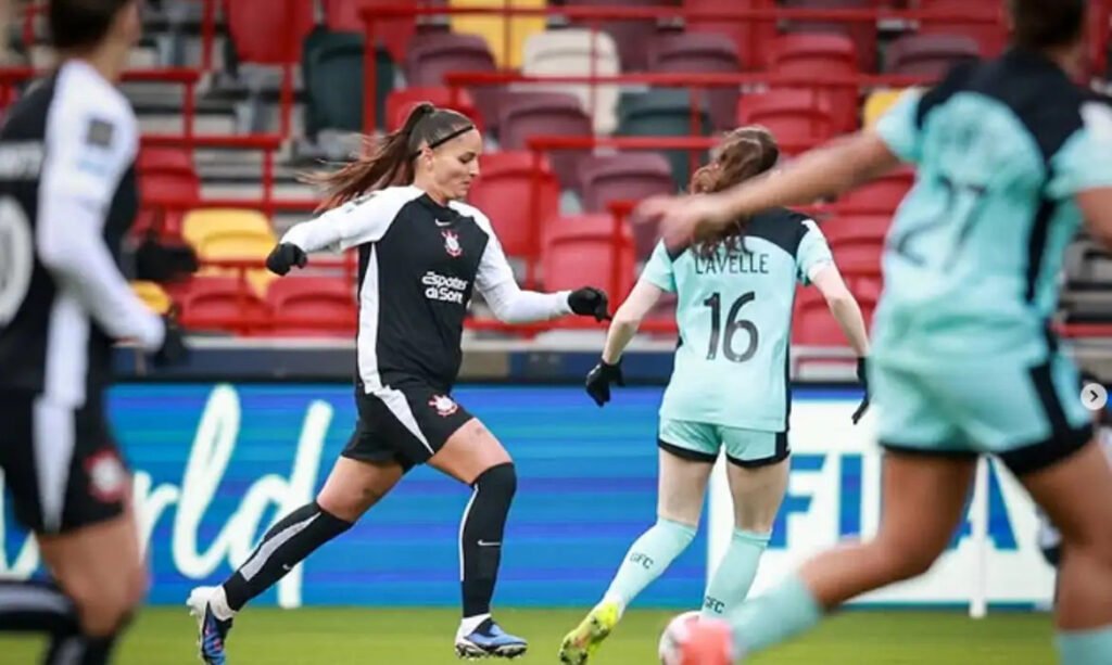 Jogadoras do Corinthians e do Liverpool disputam partida de futebol feminino em estádio com arquibancadas vermelhas.