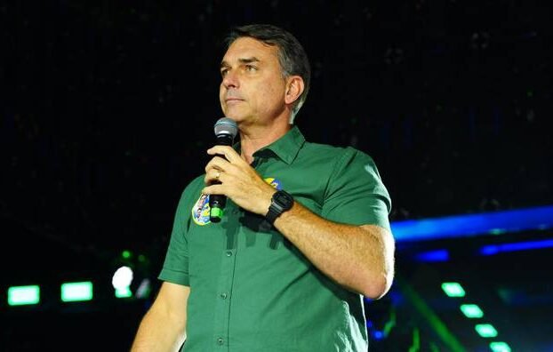 Flávio Bolsonaro veste camisa verde e segura um microfone durante discurso em evento político.