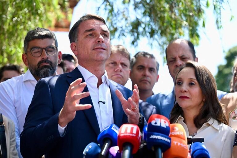 Flávio Bolsonaro fala à imprensa durante entrevista coletiva, diante de microfones e acompanhado por outras pessoas