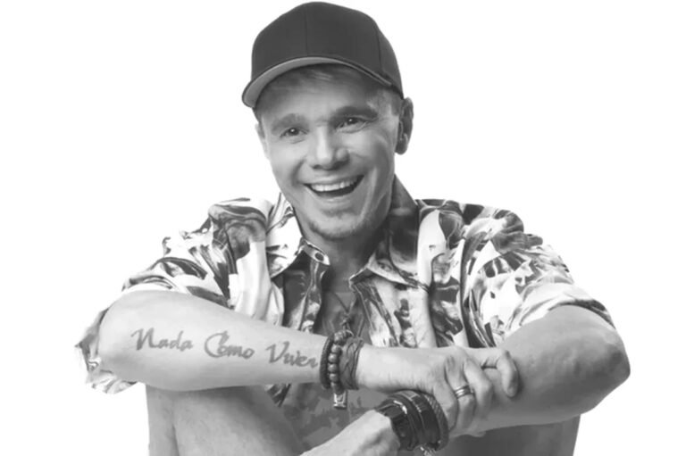 Cantor Netinho sorrindo, usando boné e camisa estampada, em fotografia em preto e branco